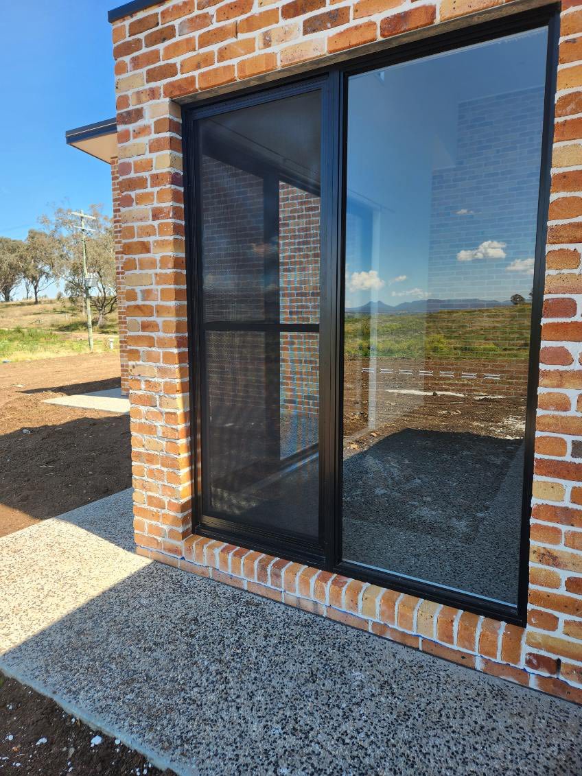 Black framed window and flyscreen on a new red brick build with Southern Downs paddocks reflected in the glass